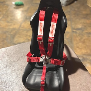 Może przedstawiać: Czarny fotel wyścigowy z czerwonymi pięciopunktowymi szelkami z logo Simpson.