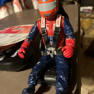 Pode incluir: Uma figura de ação de piloto de corrida vermelho e azul usando um capacete com a palavra "DUPONT" nele. A figura está usando um arnês de corrida preto com a palavra "SIMPSON" nas correias. A figura está sentada em um assento de corrida preto.