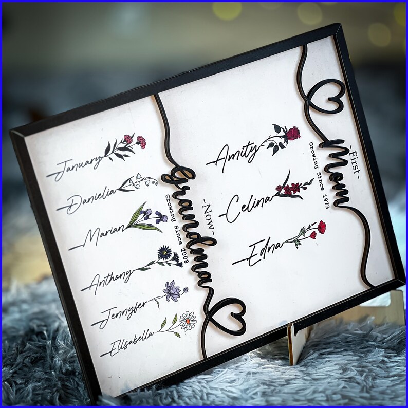 May include: A black framed sign with white background and black lettering. The sign features a decorative heart design with the words "Grandma" and "Mom" in a script font. The sign also includes a list of names and dates, including "Growing Since 2008" and "Growing Since 1973".