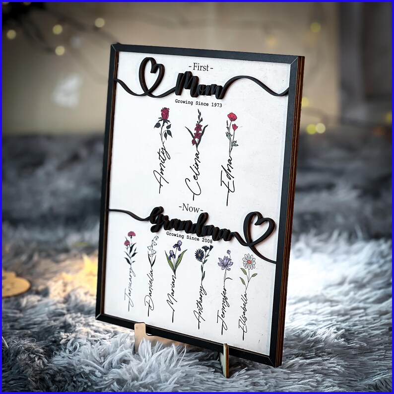 May include: A black framed wooden sign with a white background and black lettering. The sign features the words "First - Mom Growing Since 1973" and "Now - Grandma Growing Since 2008" with a heart shape next to each word. The sign also features a floral design with names written on each flower.