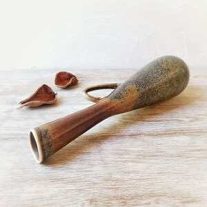May include: A handmade ceramic vase with a unique shape and a handle. The vase has a brown and tan color scheme with a textured surface. The vase is approximately 12 inches long. Decorative dried seed pods are next to the vase.