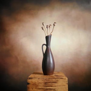 May include: A dark brown ceramic vase with a handle, holding dried seed pods. The vase is placed on a weathered wooden box. The background has a gradient of brown and tan tones, creating a warm, rustic aesthetic.