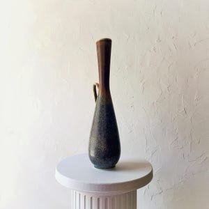May include: A tall, slender ceramic vase with a dark brown top and a gradient of green and blue hues towards the base. It features a small handle and sits on a white pedestal, showcasing its elegant, elongated form. The vase has a textured surface.