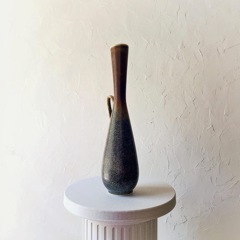 May include: A tall, slender ceramic vase with a dark brown top and a gradient of green and blue hues towards the base. It features a small handle and sits on a white pedestal, showcasing its elegant, elongated form. The vase has a textured surface.