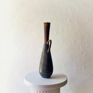 May include: A tall, slender ceramic vase with a dark brown top and a speckled, dark blue-green body. It features a gold-colored handle and sits on a white pedestal. The vase has a unique, elongated shape.