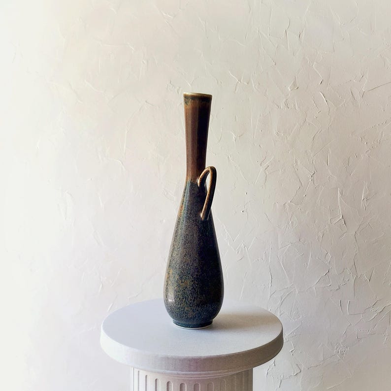 May include: A tall, slender ceramic vase with a dark brown top and a speckled, dark blue-green body. It features a gold-colored handle and sits on a white pedestal. The vase has a unique, elongated shape.