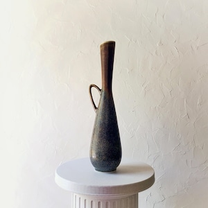 May include: A tall, slender ceramic vase with a dark brown top and a speckled blue-green body. It features a curved handle and sits on a white pedestal. The vase has a unique, elongated shape.