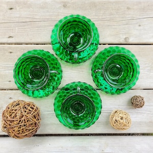 May include: Four green glass teacups and saucers with a bubbled texture. The cups and saucers are arranged in a circle on a wooden surface.