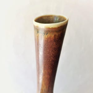 May include: A tall, ceramic vase with a flared top. The vase has a brown, textured exterior with a gradient of colors, including shades of brown, orange, and green. The rim is a creamy off-white color.