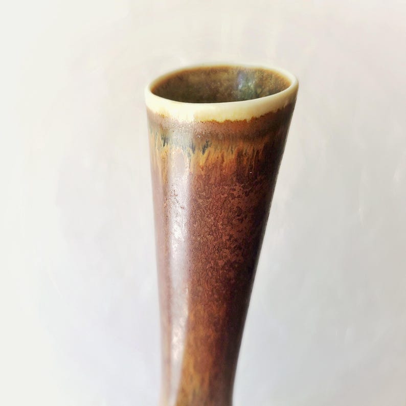 May include: A tall, ceramic vase with a flared top. The vase has a brown, textured exterior with a gradient of colors, including shades of brown, orange, and green. The rim is a creamy off-white color.