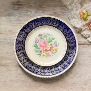 May include: Two vintage porcelain plates. The larger plate has a navy blue border with gold filigree. The smaller plate features a floral design with a pink rose and other colorful flowers. The plates are on a light wood surface.