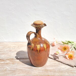 May include: A handmade ceramic jug with a cork stopper. The jug has a brown and orange glaze, a handle, and a spout. The background includes a white wall and a wooden surface. Decorative flowers are in the background.