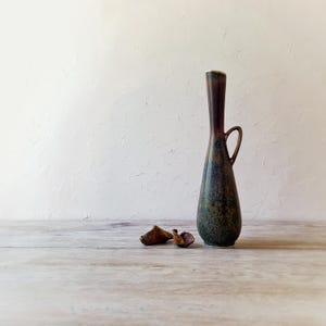 May include: A tall, slender ceramic vase with a dark brown and green speckled glaze. The vase has a small handle and a flared top. Two small, dried seed pods rest on the wooden surface in front of the vase.