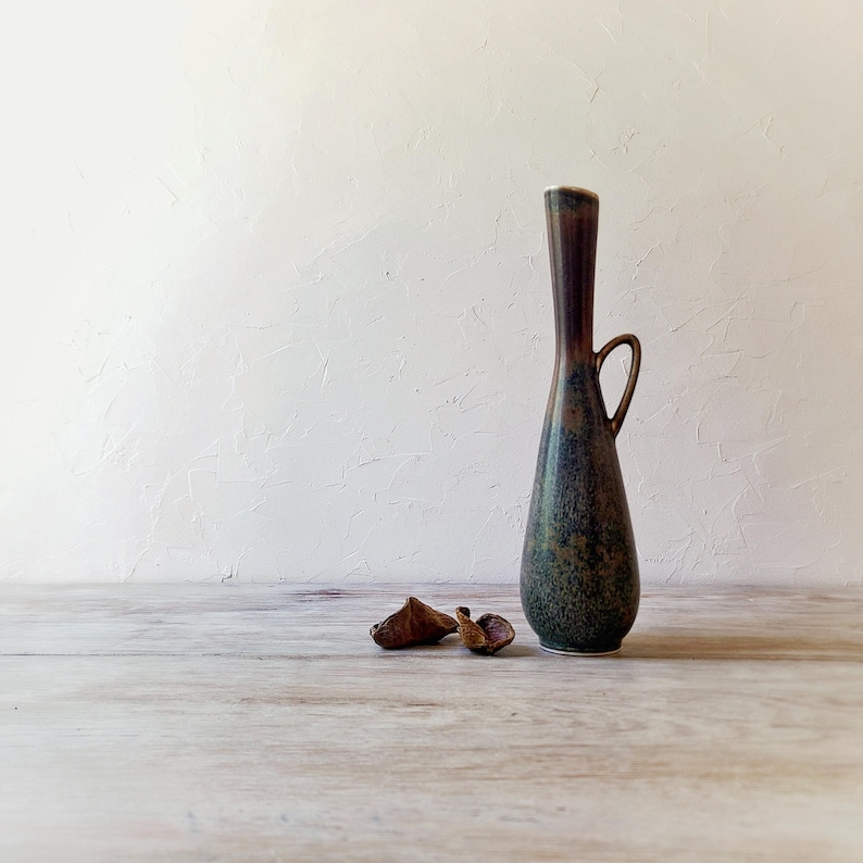 May include: A tall, slender ceramic vase with a dark brown and green speckled glaze. The vase has a small handle and a flared top. Two small, dried seed pods rest on the wooden surface in front of the vase.