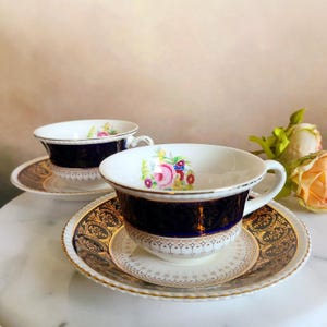 May include: Two vintage teacups and saucers with a blue and gold floral design. The cups and saucers have a white background with a dark blue band around the rim. The floral design is in shades of pink, yellow, and green. The saucers have a gold trim.