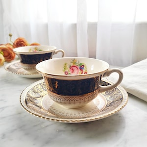 May include: Two vintage teacups and saucers. The cups have a cream-colored interior with a floral design and a dark blue exterior with gold accents. The saucers have a matching design. The set is placed on a marble surface.