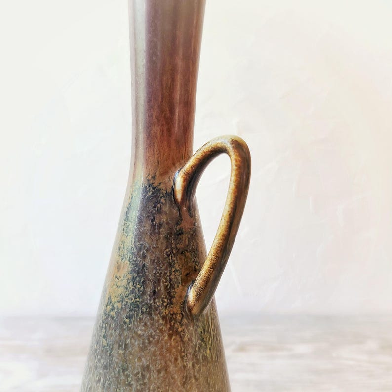 May include: A tall, brown ceramic vase with a handle. The vase has a slender neck and a wider base, with a curved handle on the side. The color transitions from a dark brown at the top to a lighter brown and green at the base. The vase has a glossy finish.