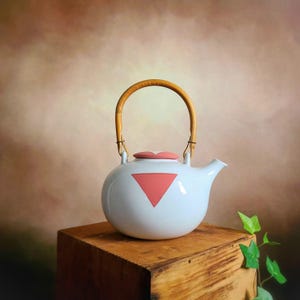 以下が含まれることがあります： ピンクの三角形のデザインと竹製のハンドルが付いた白いセラミック製のティーポット。ティーポットは木製の表面の上に置かれています