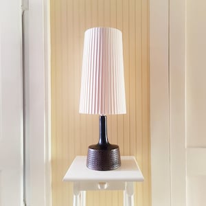 Peut inclure: Une lampe de table moderne avec un abat-jour plissé blanc et une base noire. La base présente un motif texturé. La lampe est posée sur une petite table blanche contre un mur jaune clair.
