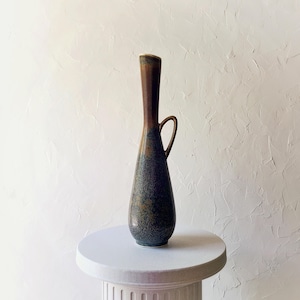 May include: A tall, slender ceramic vase with a dark brown top and a blue-green speckled body. It features a small handle and sits on a white pedestal. The vase has a unique, elongated shape and a glossy finish.