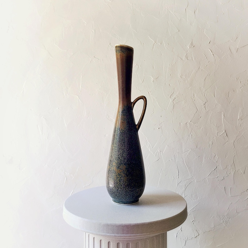 May include: A tall, slender ceramic vase with a dark brown top and a blue-green speckled body. It features a small handle and sits on a white pedestal. The vase has a unique, elongated shape and a glossy finish.