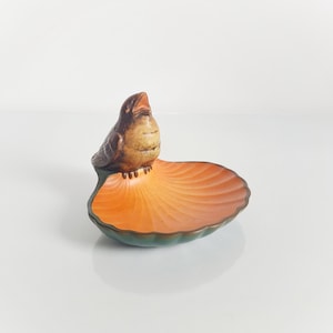 May include: A decorative ceramic dish featuring a bird perched on a leaf-shaped base. The bird is brown and tan, with an open beak. The dish has an orange interior and a green rim, creating a nature-inspired design.