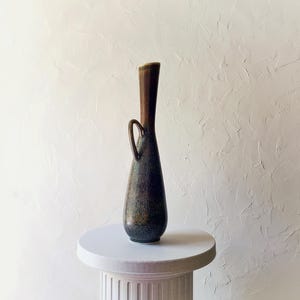 May include: A tall, slender ceramic vase with a dark, mottled glaze. The vase has a brown neck and a small handle. It is displayed on a white pedestal against a textured white background. The vase's shape is reminiscent of a vintage design.