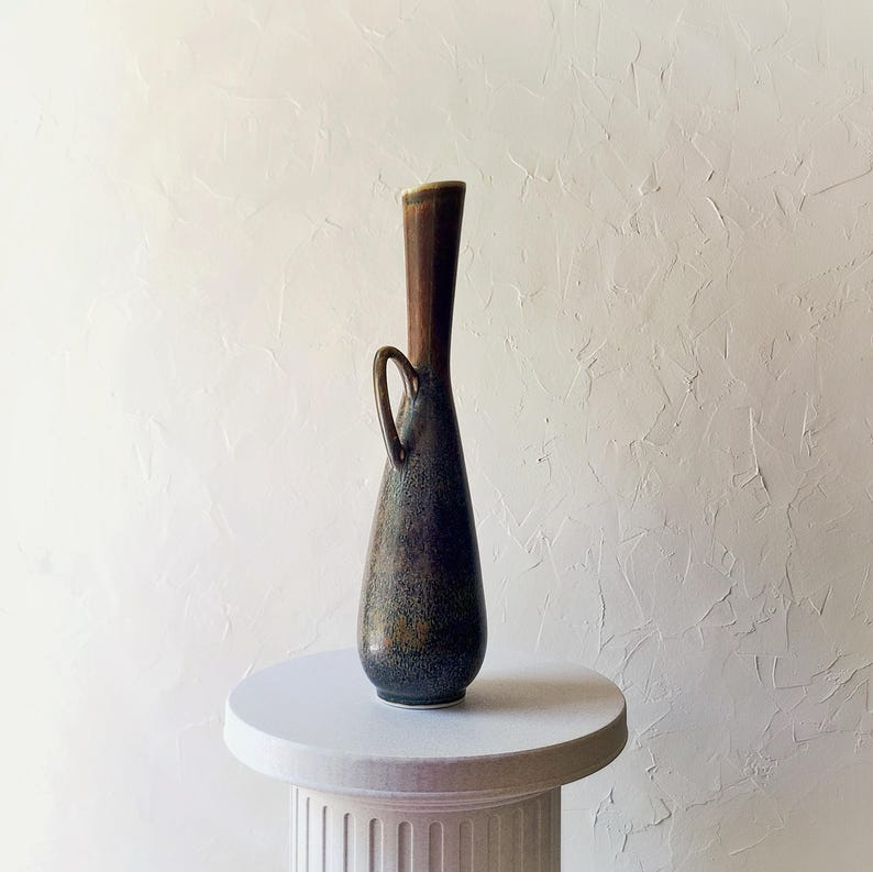 May include: A tall, slender ceramic vase with a dark, mottled glaze. The vase has a brown neck and a small handle. It is displayed on a white pedestal against a textured white background. The vase's shape is reminiscent of a vintage design.