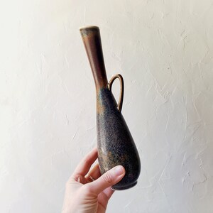 May include: A tall, slender vase with a dark brown and blue speckled finish. The vase has a long, curved neck and a small handle. The vase is held by a hand, showcasing its size and shape. The vase is likely made of ceramic or pottery.