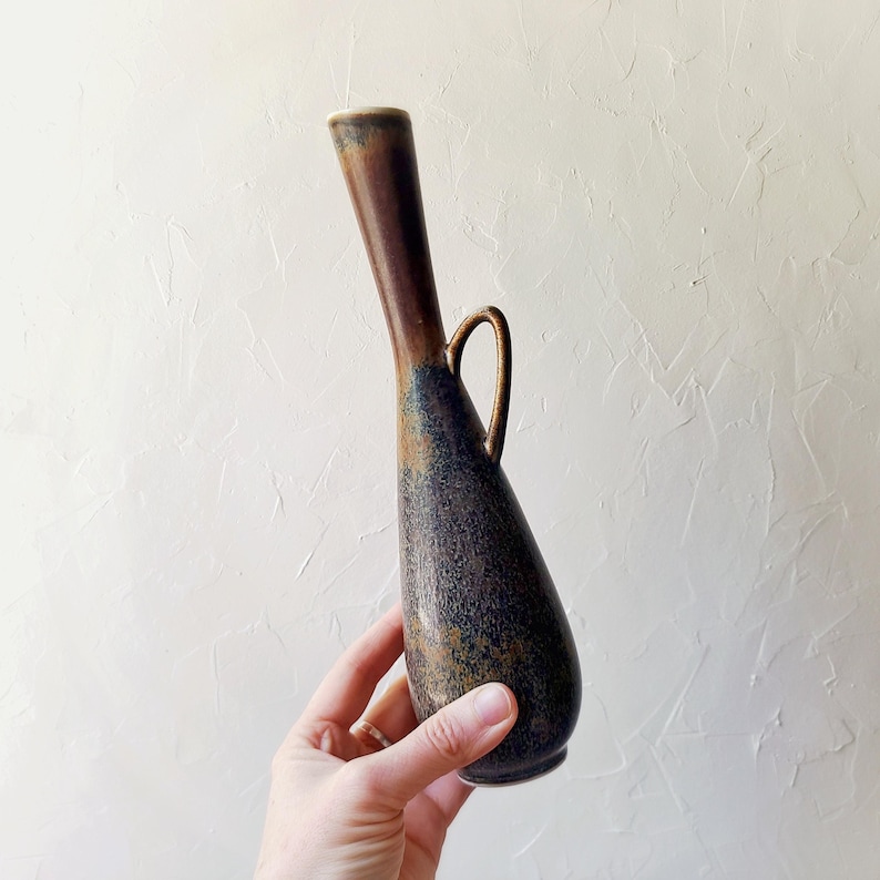 May include: A tall, slender vase with a dark brown and blue speckled finish. The vase has a long, curved neck and a small handle. The vase is held by a hand, showcasing its size and shape. The vase is likely made of ceramic or pottery.