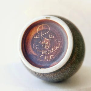 May include: A small, round, ceramic vase with a textured, dark green exterior. The base features etched text that reads "R Sweden CAF" in a brown hue, with a white rim. The vase has a simple, elegant design.