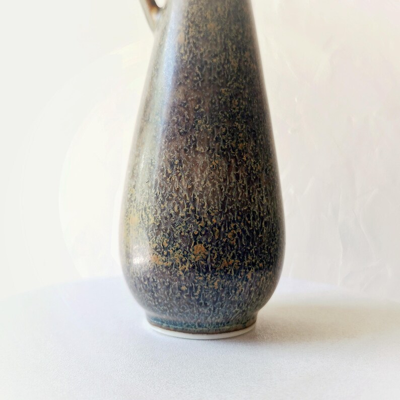 May include: A tall, textured ceramic pitcher with a handle. The pitcher has a dark blue and brown mottled glaze with gold accents. The base is white. The pitcher is likely used for decorative purposes or serving beverages.
