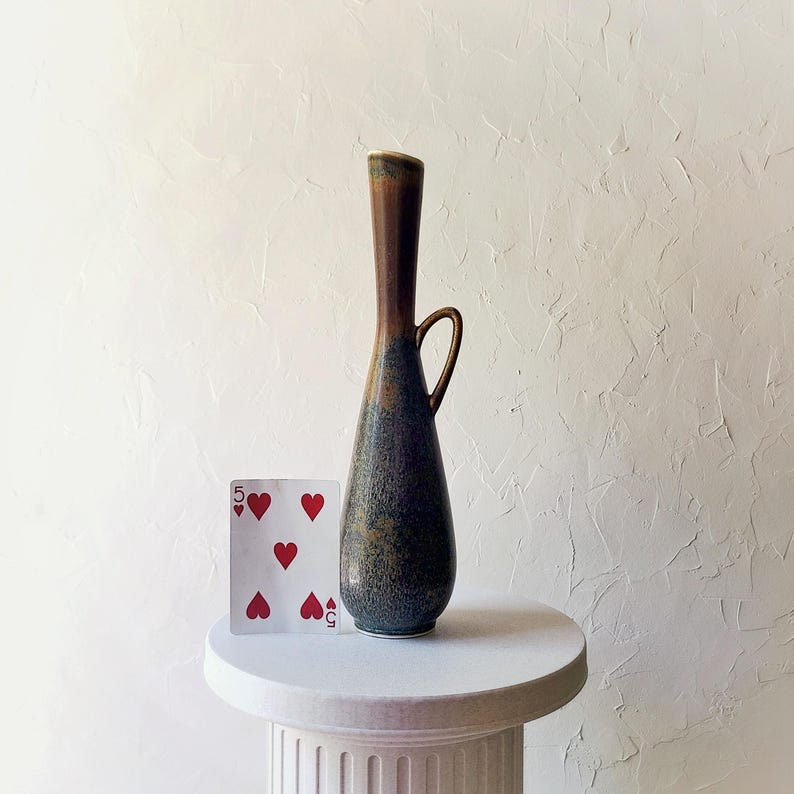 May include: A tall, slender ceramic vase with a handle, featuring a gradient of colors from brown to blue-green. A playing card with five hearts is next to the vase. The vase is on a white pedestal.