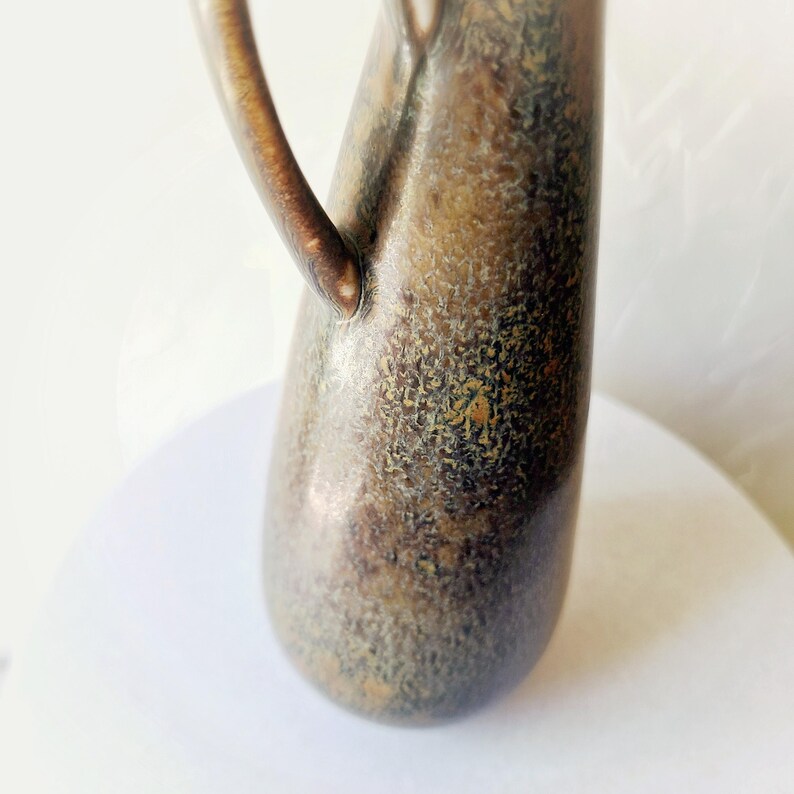 May include: A bronze-colored pitcher with a textured surface and a curved handle. The pitcher has a narrow neck and a rounded base. The handle is attached to the side of the pitcher and curves upwards. The pitcher is likely used for serving beverages.