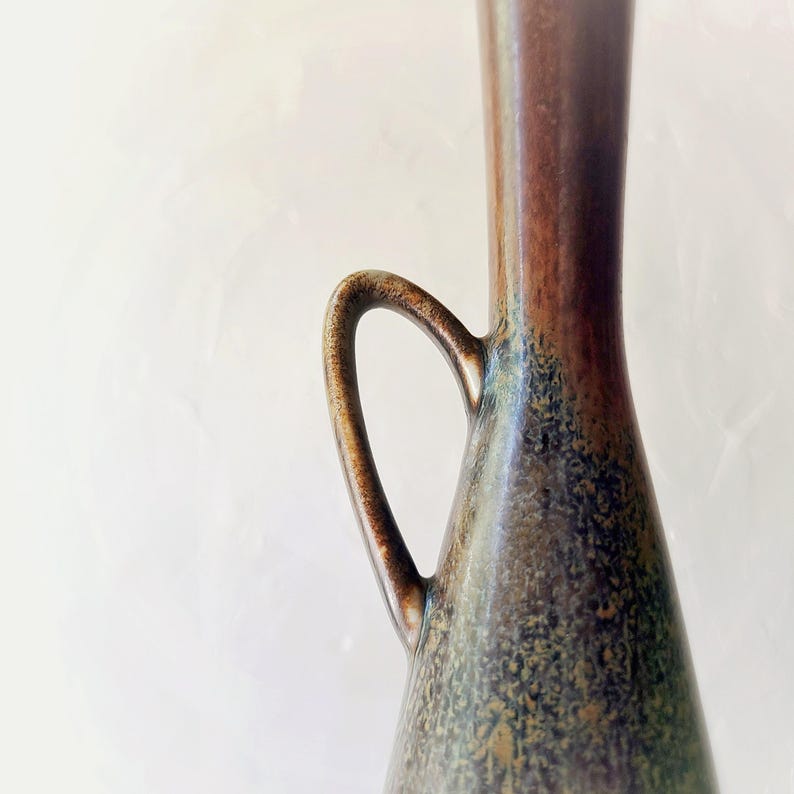 May include: A tall, slender vase with a handle, featuring a gradient of colors. The top is a dark brown, transitioning to a bronze hue, and then to a speckled green and blue at the base. The handle is curved and integrated into the vase's design.