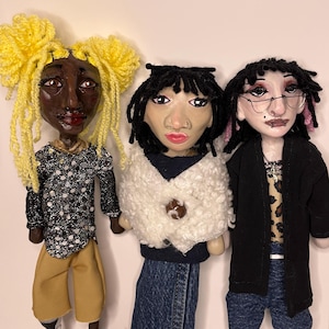 May include: Three handmade dolls with distinct features. The left doll has bright yellow yarn hair, a black and white polka dot top, and tan pants. The center doll has black hair, a white textured shawl, and a denim skirt. The right doll wears glasses, a black cardigan, and blue jeans.