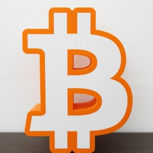 Puede incluir: Un símbolo de Bitcoin tridimensional con un marco exterior de color naranja brillante y una cara interior blanca. El diseño muestra la icónica letra B estilizada con dos trazos verticales que cruzan la parte superior e inferior, creando una estructura sólida.