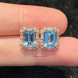 May include: A pair of stud earrings featuring rectangular emerald-cut light blue center stones surrounded by a halo of small round clear stones set in a gold-toned metal frame. The earrings are positioned side by side against a dark background.