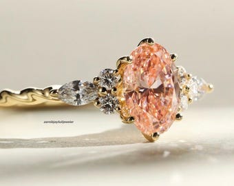 May include: A gold ring featuring a large oval-cut peach-colored center stone, flanked by smaller marquise and round-cut clear stones on each side. The band has a wavy, textured design. The text zarnibjoyfulljeweler is visible on the band.