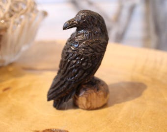 May include: A dark brown, hand-carved wooden raven figurine with a glossy finish. The bird is depicted in a standing position, showcasing detailed feather textures and a sharp beak, perched atop a small, rounded, light-coloured wooden base.
