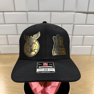 May include: Black trucker hat with a gold eagle and "AMOR ETERNO" patch. The hat is adjustable and has a Richardson 112 label.