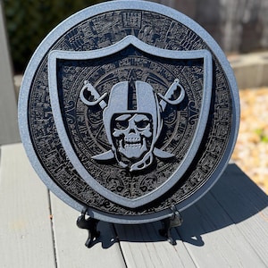 May include: A round, black and silver decorative plaque featuring the Raiders logo. The logo includes a skull wearing a football helmet, crossed swords, and a shield. The plaque is displayed on a black stand.