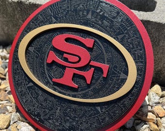 May include: A round, decorative sign featuring the San Francisco 49ers logo. The sign has a red border, a black center with an embossed pattern, and a gold oval surrounding the red "SF" logo. The sign is displayed outdoors on a bed of rocks.