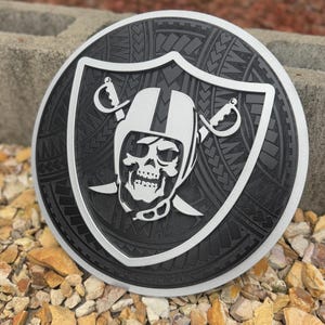 May include: A round, black and silver decorative emblem featuring a skull wearing a football helmet with crossed swords. The emblem has a tribal pattern around the edge and is set against a stone background.