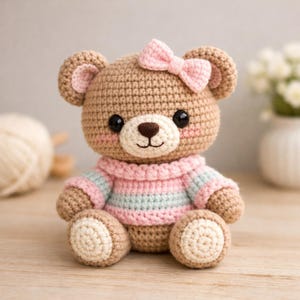 Puede incluir: Un oso de peluche de ganchillo hecho a mano con un lazo rosa, que lleva un suéter a rayas rosas y azules. El oso tiene ojos negros, una nariz marrón y mejillas rosadas. El oso está sentado sobre una superficie de madera.
