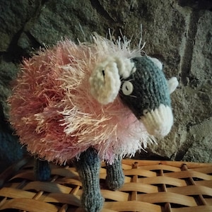 May include: A pink and white fluffy knitted sheep with grey legs and a white face. The sheep is sitting on a woven wicker basket.