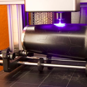 May include: A black cylindrical object is being engraved with a laser. The laser is emitting a bright purple beam of light. The object is being rotated on a metal platform.