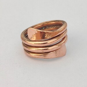 Op de afbeelding: Een koperen ring met een uniek, gedraaid ontwerp. De ring is gemaakt van meerdere koperen banden die met elkaar zijn verweven en gehamerd om een ​​getextureerd oppervlak te creëren.