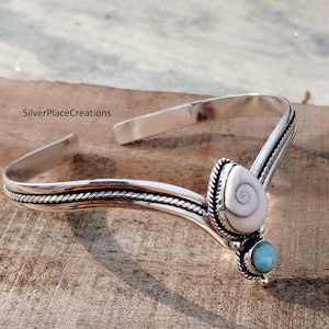 May include: A silver-toned bracelet with a white seashell and a small, round, light blue stone. The bracelet has a twisted rope design along the edges and is set against a wooden surface. The text "SilverPlaceCreations" is visible.