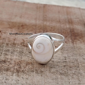 Può includere: Un anello d'argento con una grande pietra bianca ovale con un motivo a spirale. L'anello ha un design a gambo diviso. Il testo "SilverPlaceCreations" è visibile nell'angolo in alto a sinistra.